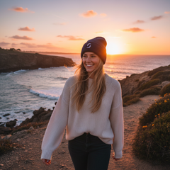 Woman standing on the coast wearing navy blue beanie. with a small logo on it.