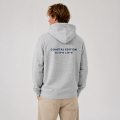 A man standing with a "coastal edition" hoodie against a grey background.