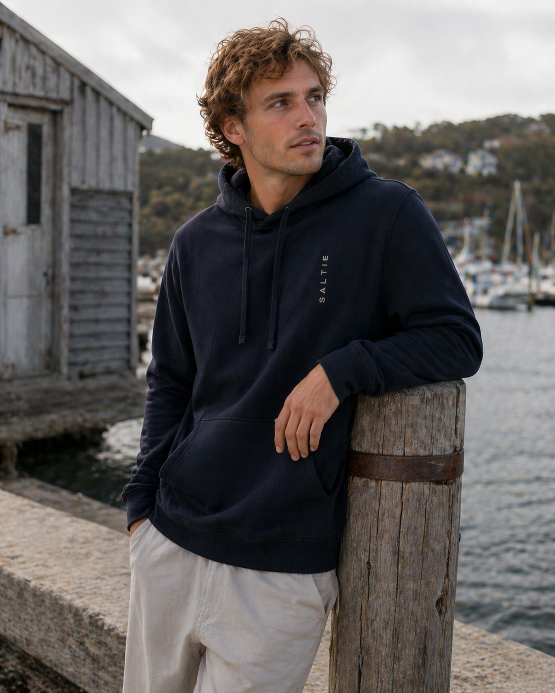 Man wearing a navy hoodie with a logo, standing by a wooden post near a marina.