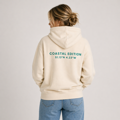 A woman facing away from the camera wearing a Biege hoodie. The woman is against a white background and the text reads "coastal editions".