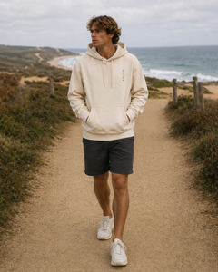 A man wearing a biege hoodie walking along the coast towards the camera. the hoodie has a small logo that reads "saltie".