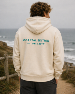 A man wearing a beige hoodie on the coast with the text "coastal edition" on the back of it.