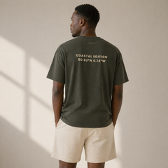 Man wearing a green t-shirt with 'Coastal Edition' text on the back, standing against a beige wall.