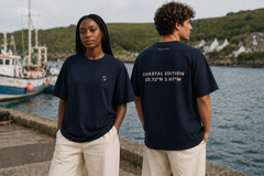 Two people wearing navy blue t-shirts with coastal design and coordinates, standing by a waterfront.