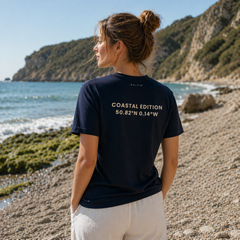 Woman standing on a beach wearing a black t-shirt with coordinates printed on the back, looking out towards the ocean.
