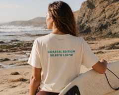 Person holding a surfboard on a beach wearing a t-shirt with coastal coordinates.